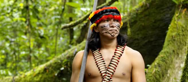An indigenous man wearing feathers and a painted face standing in the forest with a blowpipe