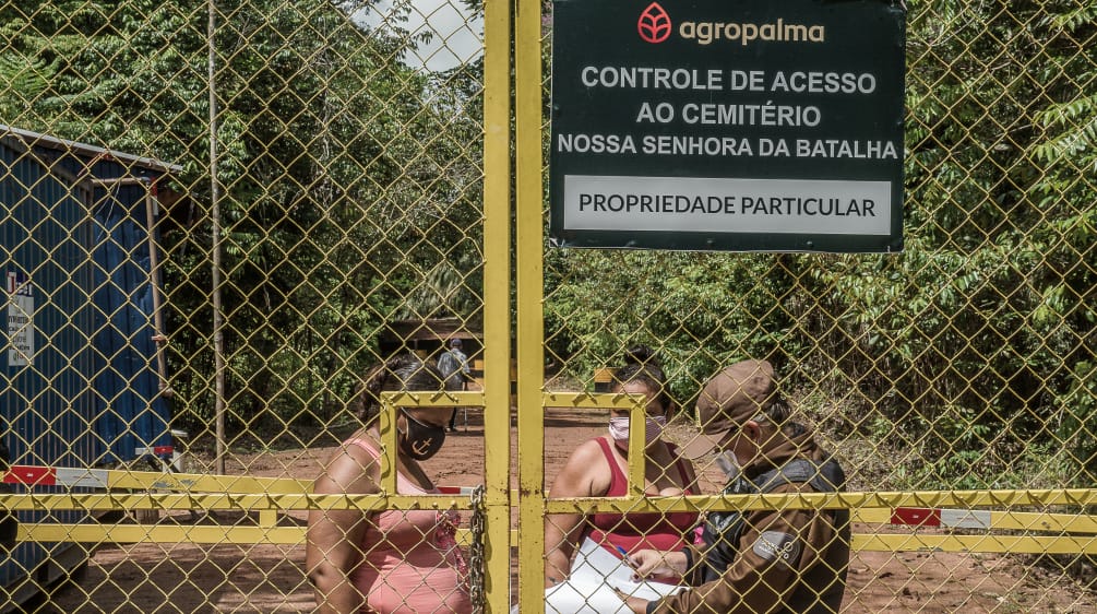Two women behind a high barred gate are being checked by a guard. Inscription on the sign: Agropalma. Access control for Nossa Senhora da Batalha cemetery. Private property