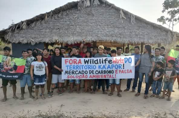 A group of Indigenous people with protest banners in front of a hut thatched with palm fronds