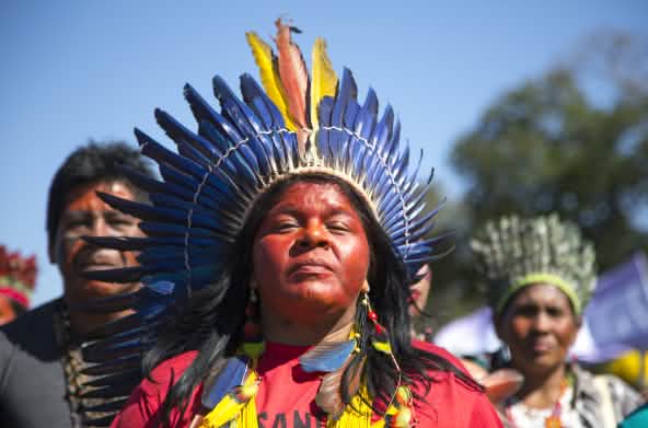 An indigenous woman adorned with a feathered crown in front of two other indigenous people