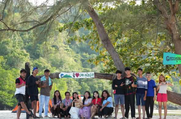 A group of young people sitting and standing in front of a tree on the beach