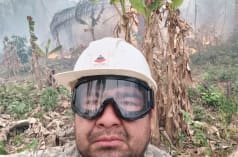 A man looks into the camera wearing a helmet and goggles while bushes burn and smoke rises in the background