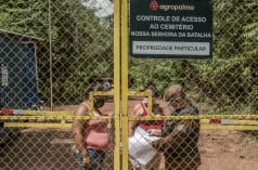Two women behind a high barred gate are being checked by a guard. Inscription on the sign: Agropalma. Access control for Nossa Senhora da Batalha cemetery. Private property