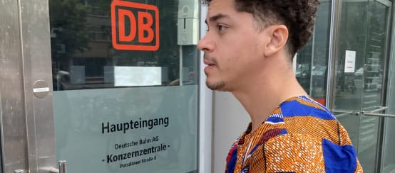 A young man stands with an envelope in front of a glass door with the inscription DB Main Entrance, Corporate Headquarters