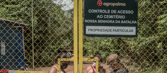 Two women behind a high barred gate are being checked by a guard. Inscription on the sign: Agropalma. Access control for Nossa Senhora da Batalha cemetery. Private property