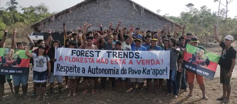 Ka'apor protesting carbon traders in their territory. Banner: Forest Trends. The rainforest is not for sale. Respect the autonomy of the Ka'apor people! A group of Indigenous people with protest banners in front of a hut covered with palm fronds