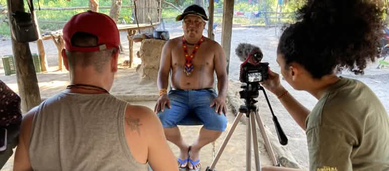 The Intercept journalists interviewing a member of the Council of the Ka'apor Two journalists in the foreground record a video interview with an Indigenous leader (in the background) using a tripod-mounted camera