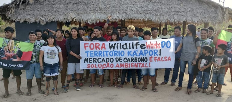 Ka’apor protest against carbon traders in their territory. Inscription on the banner: Wildlife Works out of Ka’apor territory! The carbon market is a false environmental solution! A group of Indigenous people with protest banners in front of a hut thatched with palm fronds