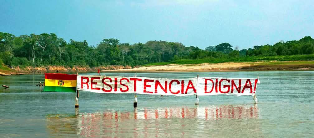 Inhabitants of the TIPNIS national park are protesting a planned road through the rainforest. Protest banner on a river in the Bolivian rainforest: the slogan “Resistance Means Dignity” stands next to the Bolivian flag.