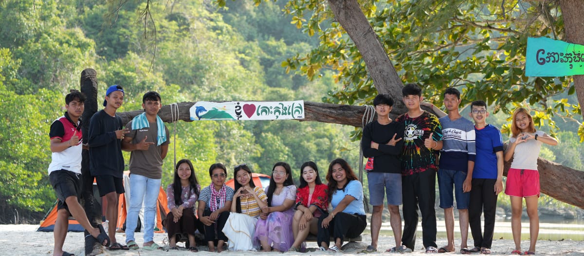 A group of young people sitting and standing in front of a tree on the beach