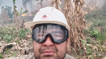 A man looks into the camera wearing a helmet and goggles while bushes burn and smoke rises in the background