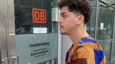 A young man stands with an envelope in front of a glass door with the inscription DB Main Entrance, Corporate Headquarters