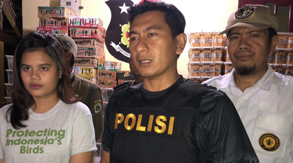 Young woman wearing Flight t-shirt and police officer in a warehouse with crates of birds